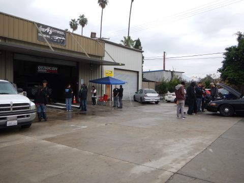 2003 Ford Crown Victoria - white Dodge Ram pickup and several sedans sit in a damp auto shop lot as people gather near a blue pop-up tent.