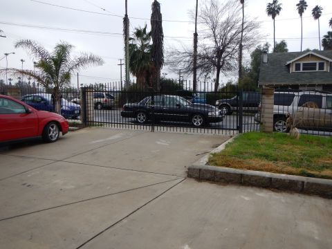 2003 Ford Crown Victoria - red coupe and a dark sedan sit on a concrete lot behind a black metal fence, with palm trees and a bungalow in the background.
