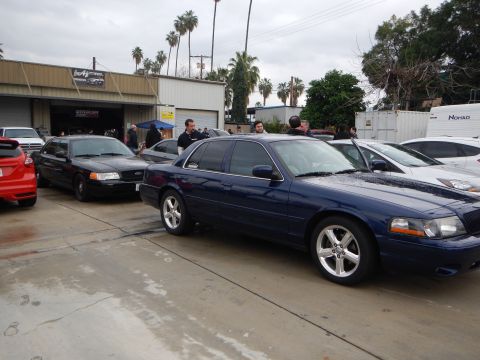 2003 Ford Crown Victoria - dark blue Mercury sedan with polished alloy wheels is parked among other cars in a busy auto shop lot.