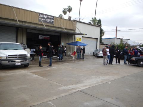 2003 Ford Crown Victoria - People and a small dog gather in a wet auto shop lot beside a white pickup, a blue tent, and parked cars under a cloudy sky.