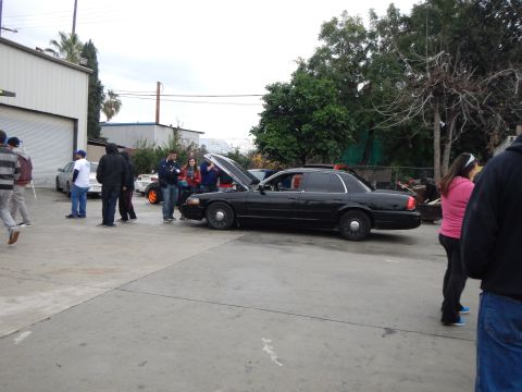 2003 Ford Crown Victoria - People stand around a black sedan with its hood up in an outdoor lot beside an industrial building and trees.