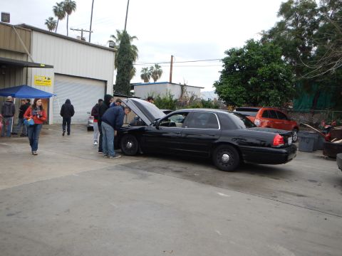 2003 Ford Crown Victoria - black Ford sedan sits with its hood up as people inspect the engine in front of an auto shop, with an orange SUV nearby.
