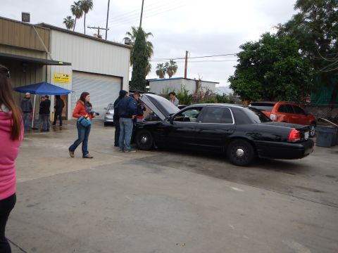 2003 Ford Crown Victoria - black sedan with its hood raised is parked outside an auto shop while people stand around it under an overcast sky.