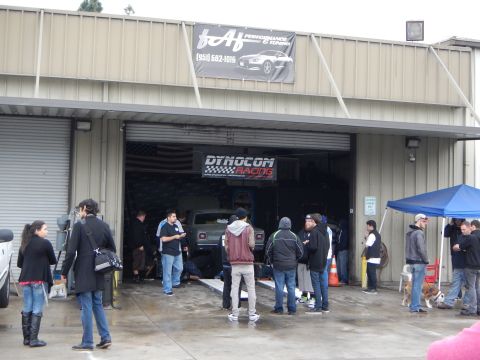 2003 Ford Crown Victoria - People gather outside an open auto shop bay displaying a Dynocom Racing banner, with a truck on a dyno visible inside.
