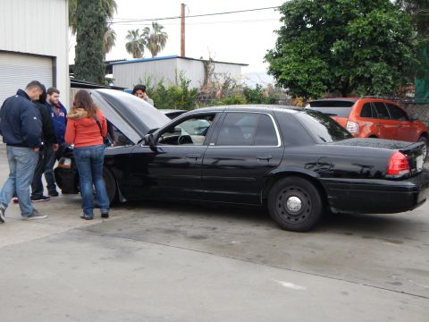 2003 Ford Crown Victoria - black Ford sedan with steel wheels and an open hood is examined by several people in an outdoor lot.