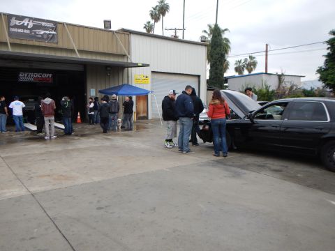 2003 Ford Crown Victoria - People mill about a wet auto shop lot with a blue tent, a white pickup, and a black sedan with its hood raised.