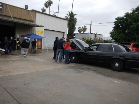 2003 Ford Crown Victoria - black sedan with its hood up is surrounded by people near a blue pop-up tent outside an industrial auto shop.