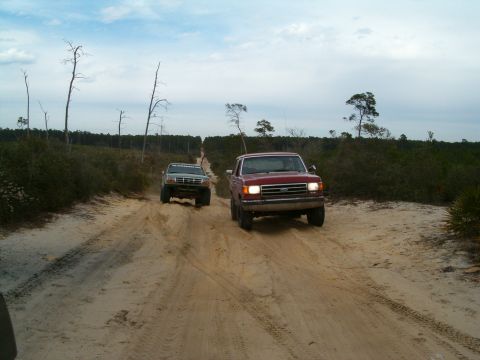 1991 Ford Bronco - A red 1991 Ford Bronco leads a green truck down a sandy track through a wooded area.