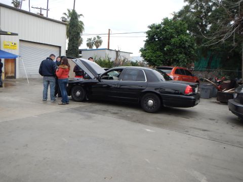 2003 Ford Crown Victoria - black sedan with its hood open sits in a concrete lot as a few people inspect it, with an orange SUV parked behind.