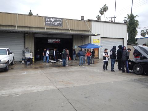 2003 Ford Crown Victoria - crowd stands in a wet lot outside an auto shop where a car sits on a lift inside the open garage bay.