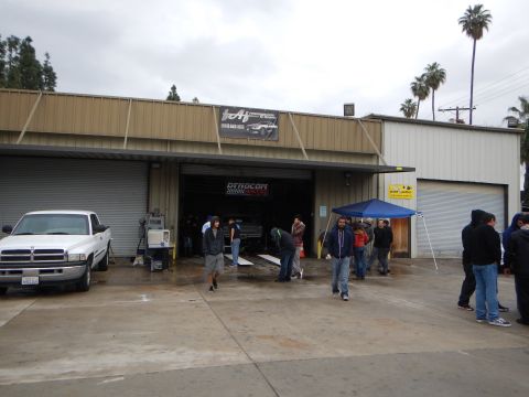 2003 Ford Crown Victoria - white Dodge Ram pickup is parked beside a crowd gathered outside an open auto shop garage with a Dynocom banner inside.