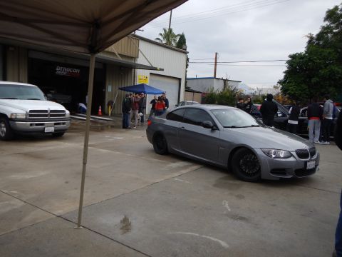 2003 Ford Crown Victoria - gray BMW coupe with black alloy wheels is parked in a wet auto shop lot, framed by a tent canopy and onlookers.