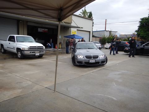 2003 Ford Crown Victoria - gray BMW coupe with angel-eye headlights faces forward in an auto shop lot next to a white pickup, with people standing nearby.