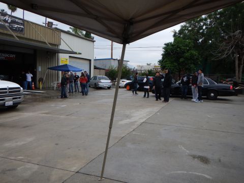2003 Ford Crown Victoria - wide shot of a damp auto shop lot shows several cars, a white pickup, and people gathered near a blue tent under a gray sky.