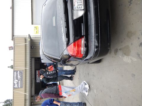 2003 Ford Crown Victoria - rear corner of a black sedan shows a personalized 'EVIL' license plate as people stand near an auto shop entrance.