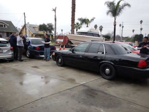 2003 Ford Crown Victoria - black Ford sedan is parked on a wet lot next to a boat on a trailer and several other cars, with palm trees and houses behind.
