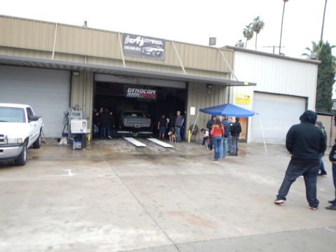 2003 Ford Crown Victoria - People gather outside an auto shop where a car sits raised on a lift inside the open garage bay beside a white pickup and blue tent.