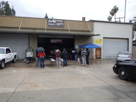 2003 Ford Crown Victoria - crowd stands under umbrellas outside a tan metal shop building with an open garage bay on an overcast, rainy day.
