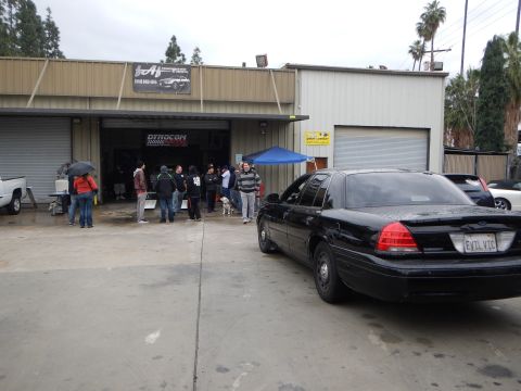 2003 Ford Crown Victoria - black four-door sedan sits in a wet lot beside a tan shop where people gather near an open garage bay under a blue canopy.