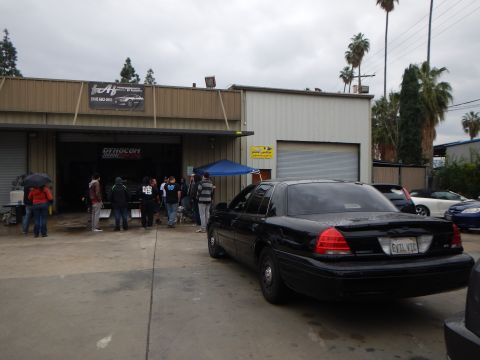 2003 Ford Crown Victoria - black sedan is parked on wet pavement outside a tan metal shop as people gather around the open garage bay.