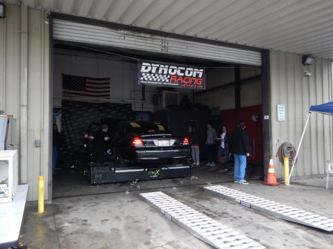 2003 Ford Crown Victoria - black sedan sits on a dynamometer inside a garage beneath a Dynocom Racing banner and a US flag, with onlookers nearby.