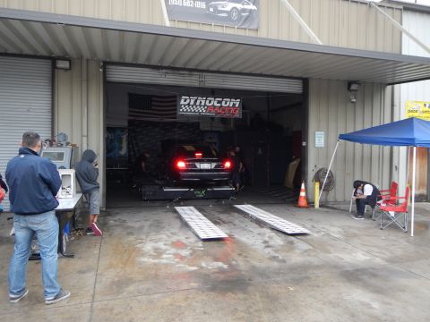 2003 Ford Crown Victoria - black sedan runs on a dyno with red taillights lit, inside a garage marked Dynocom Racing, ramps laid out and onlookers watching.