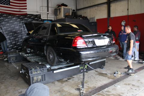 2003 Ford Crown Victoria - black Ford Crown Victoria sits on a dynamometer with its hood up inside a garage, a US flag on the wall and people standing alongside.