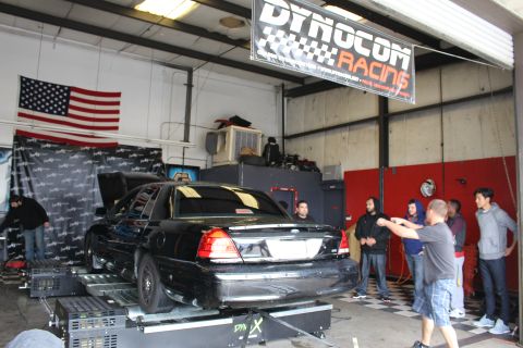 2003 Ford Crown Victoria - black sedan sits on a dyno beneath a Dynocom Racing banner and a US flag while a group of people watch from the side.