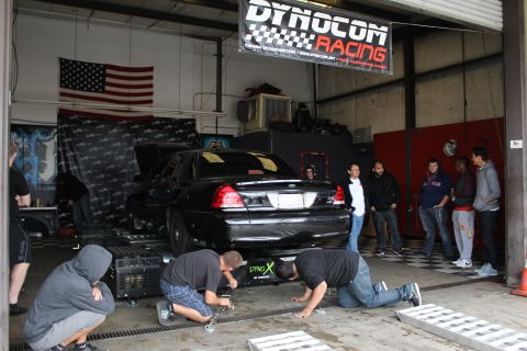 2003 Ford Crown Victoria - Mechanics kneel and work at the front of a black sedan on a dyno beneath a Dynocom Racing banner as a crowd looks on.