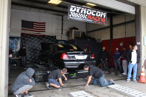2003 Ford Crown Victoria - black Ford sedan sits on a dyno while three people kneel at its front, beneath a Dynocom Racing banner with onlookers nearby.