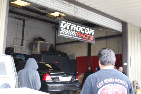 2003 Ford Crown Victoria - Looking into a garage bay at a black Ford sedan beneath a Dynocom Racing banner, with a man in a graphic shirt in the foreground.