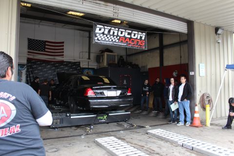 2003 Ford Crown Victoria - black sedan sits on a dyno inside an open garage bay beneath a Dynocom Racing banner, with ramps and people in front.