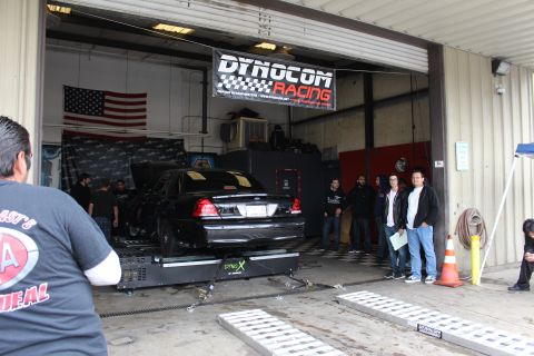 2003 Ford Crown Victoria - black sedan rests on a dyno inside an open garage bay under a Dynocom Racing banner as several people stand watching outside.