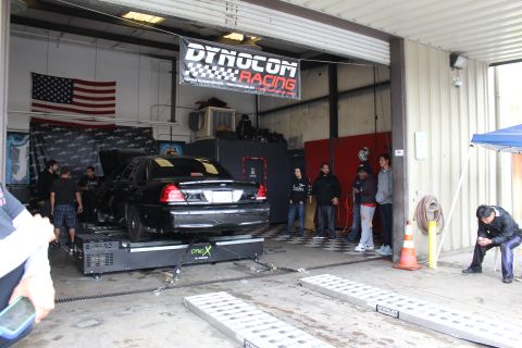 2003 Ford Crown Victoria - black Ford sedan sits on a dyno inside a garage bay beneath a Dynocom Racing banner, with onlookers and an orange traffic cone nearby.