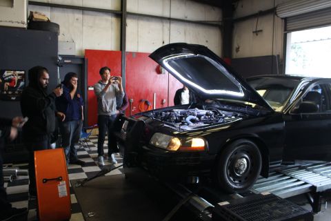 2003 Ford Crown Victoria - black Ford sedan sits on a dyno with its hood raised, showing the engine bay, while several people watch and take photos.