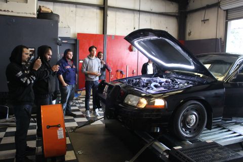 2003 Ford Crown Victoria - black Ford sedan sits on a dyno with its hood up while a man addresses a group of onlookers in the garage.