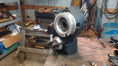 1995 Ford Bronco - A black transfer case with a silver bolted flange sits on a cluttered workbench in a garage setting.