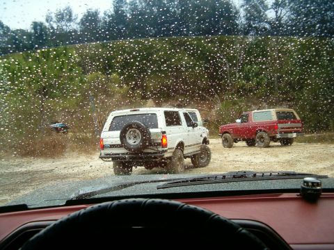 1991 Ford Bronco - Two Ford Broncos drive on a dirt road in wet weather, seen through a rain-streaked windshield.