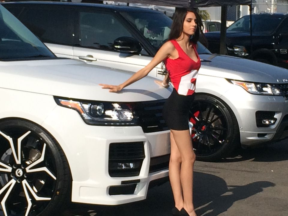 Woman in red top and black skirt leans on white Range Rover; silver Range Rover behind