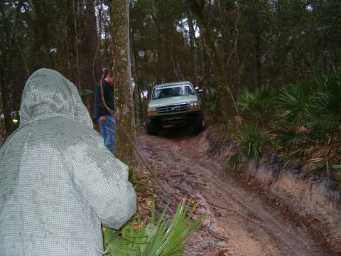 1991 Ford Bronco - 1991 Ford Bronco tackling a challenging muddy off-road trail surrounded by trees.
