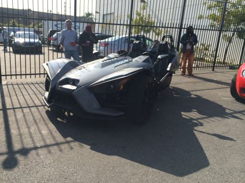 1968 Ford Mustang - A Polaris Slingshot is parked behind a fence, with other vehicles and people visible.