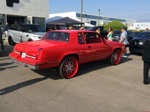 1968 Ford Mustang - A red coupe with oversized custom wheels is parked outdoors, with other vehicles and people in the background.