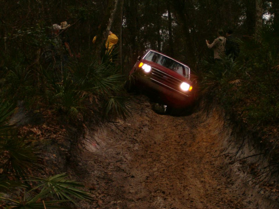 1991 Ford Bronco driving down a muddy off-road trail with trees and brush on either side