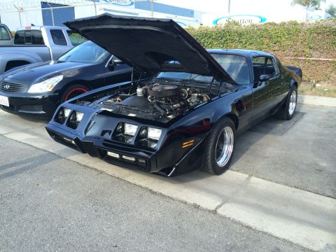1968 Ford Mustang - A black 1977 Pontiac Firebird Trans Am is parked with its hood up, revealing the engine bay.
