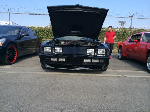 1968 Ford Mustang - Black 1977 Pontiac Firebird Trans Am with hood open, parked at an outdoor event.
