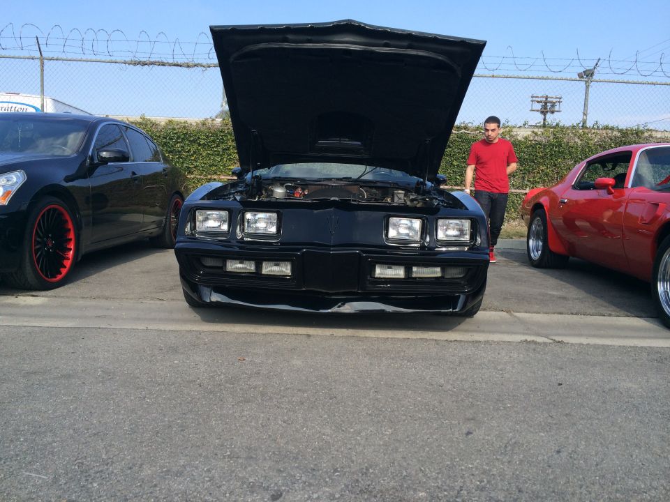 Front view of a black 1977 Pontiac Firebird Trans Am with hood open, parked next to a red Firebird and a black sedan