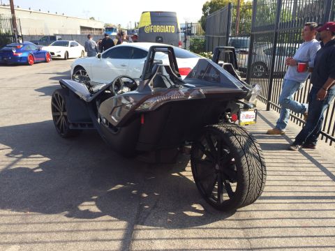 1968 Ford Mustang - A three-wheeled autocycle is parked next to other vehicles, with people standing nearby.
