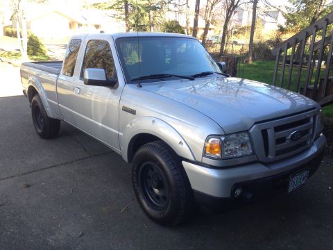 2010 Ford Ranger - This silver Ford Ranger pickup truck is shown from a 3/4 front angle, parked on a driveway with trees and a wooden fence in the background.