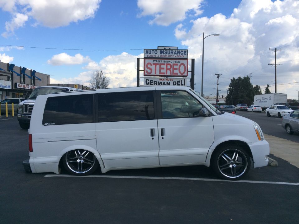 White minivan with custom wheels parked in a lot. Signage for auto insurance, stereo, and German deli visible