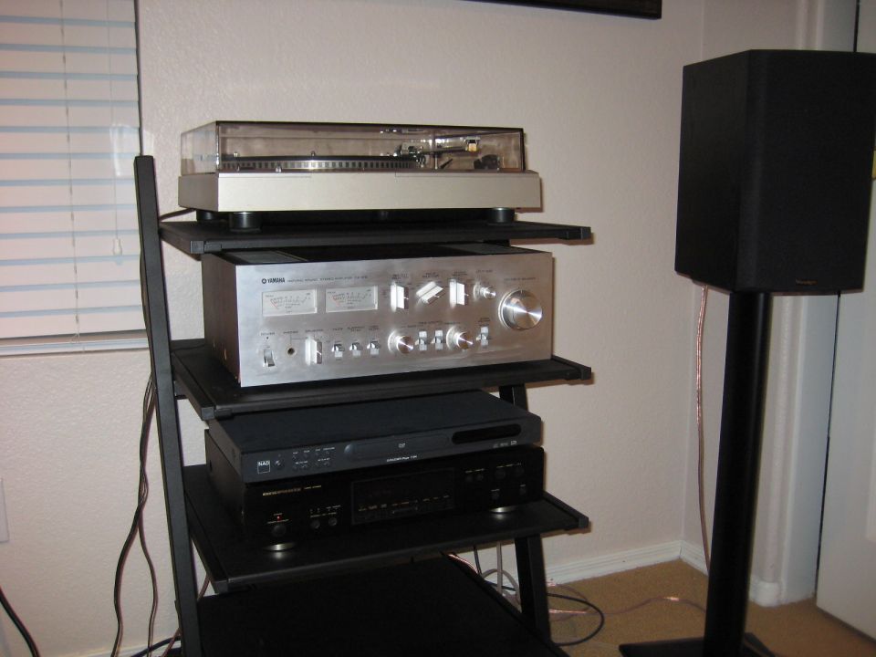 Stereo equipment rack holding a turntable, silver amplifier, and black CD player, with a speaker on a stand to the right